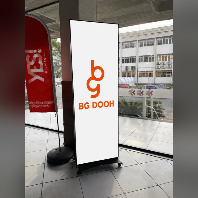 Totem de LED vertical dentro do Shopping Unisul, em área de circulação com piso claro e vidros para a rua.